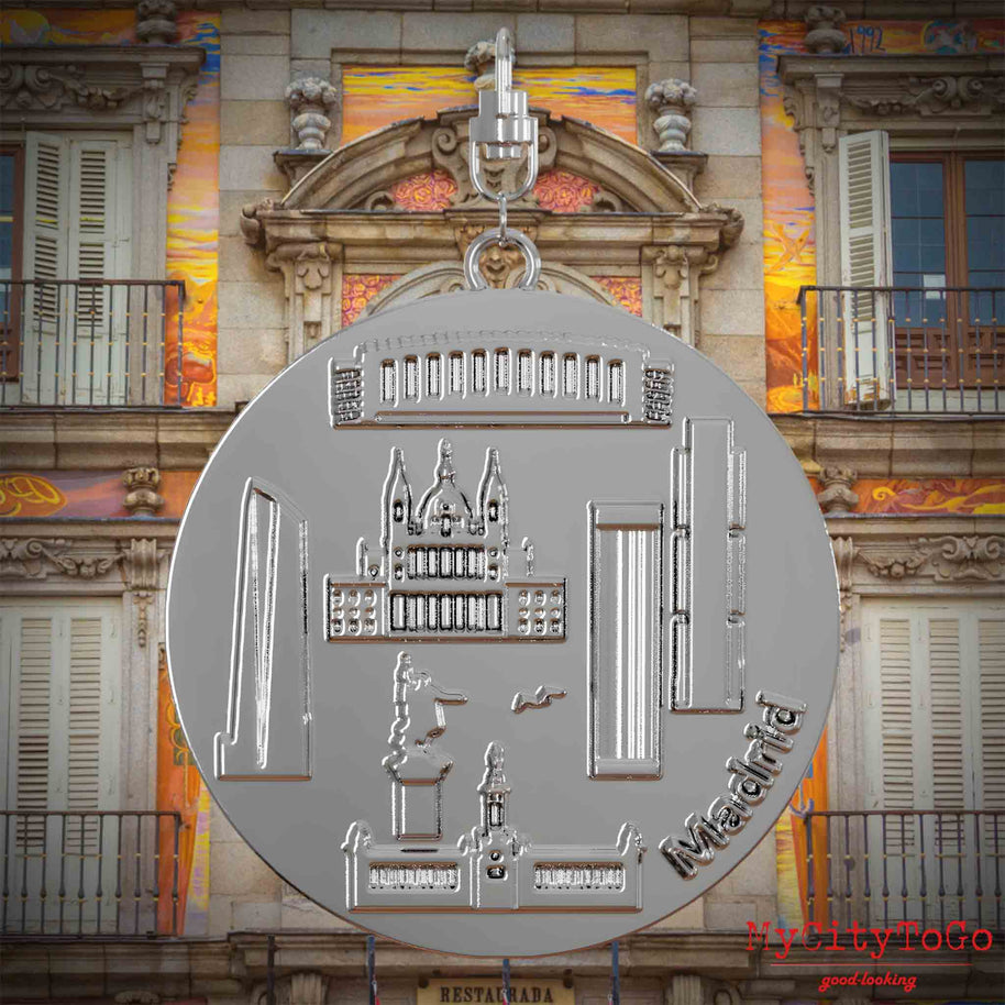 Halskette mit Drehwirbel und Rückseite eines silbernen Schmuckanhängers, auf dem Reliefs bekannter Sehenswürdigkeiten der Stadt Madrid abgebildet sind. Den Hintergrund bildet eine Ansicht der Stadt Madrid.
