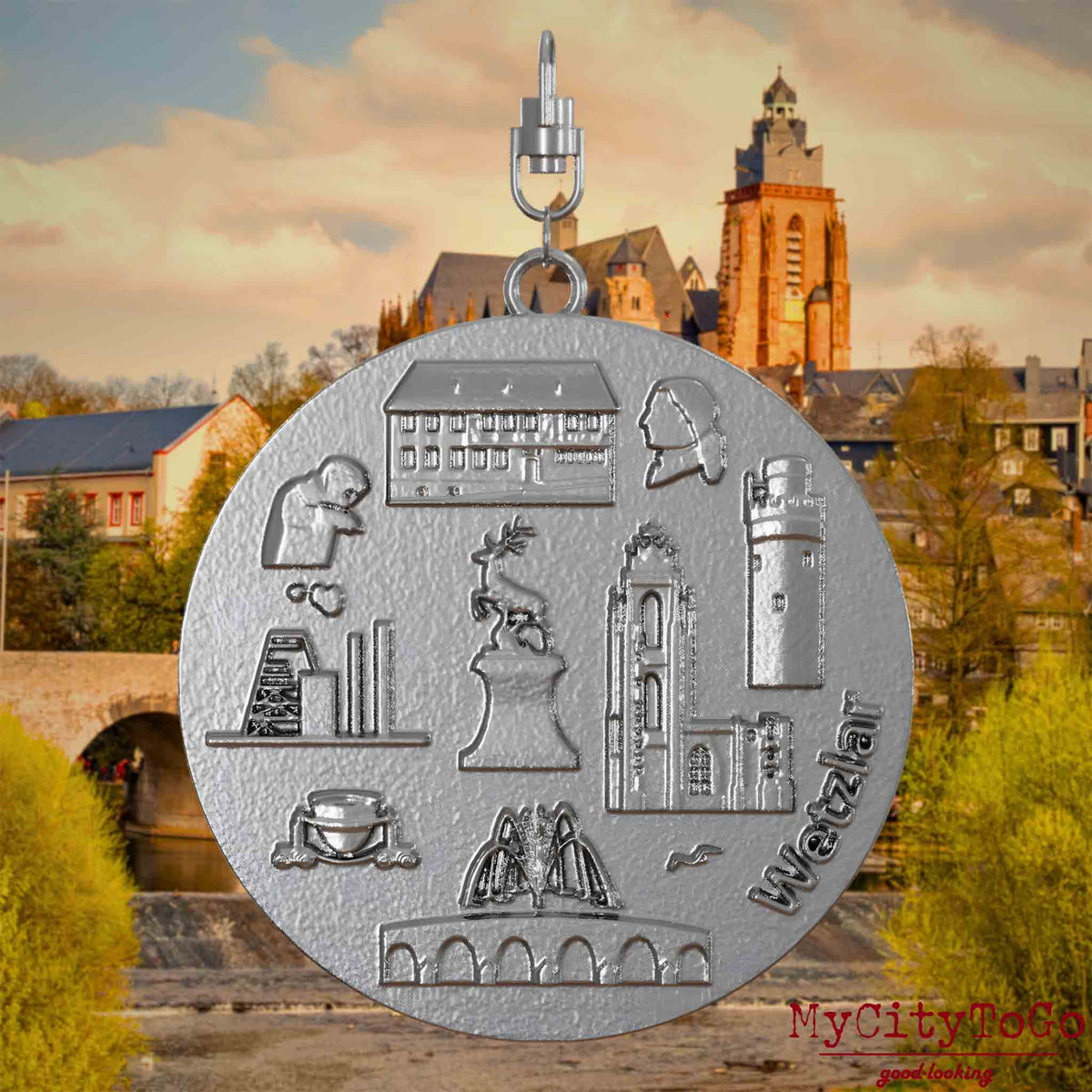 Halskette mit Drehwirbel und Rückseite eines Schmuckanhängers aus Edelstahl, auf dem Reliefs bekannter Sehenswürdigkeiten der Stadt Wetzlar abgebildet sind. Den Hintergrund bildet eine Ansicht der Stadt Wetzlar.
