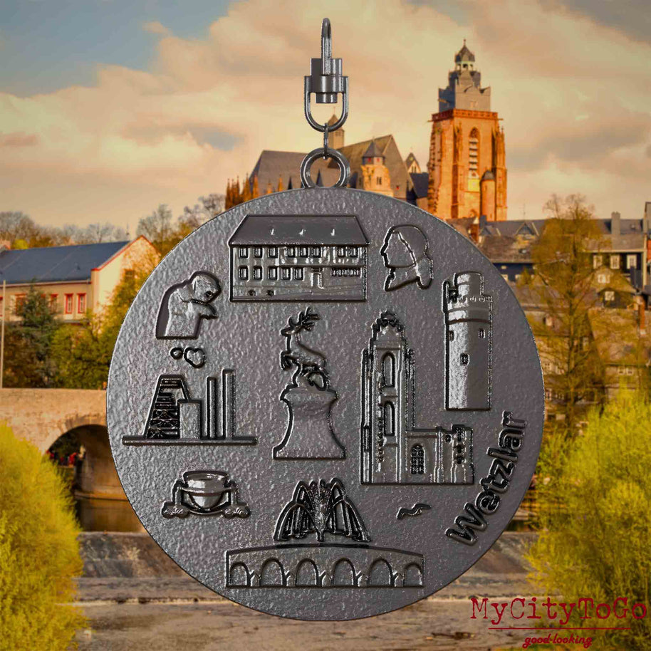 Halskette mit Drehwirbel und Rückseite eines Schmuckanhängers aus Titan, auf dem Reliefs bekannter Sehenswürdigkeiten der Stadt Wetzlar abgebildet sind. Den Hintergrund bildet eine Ansicht der Stadt Wetzlar.
