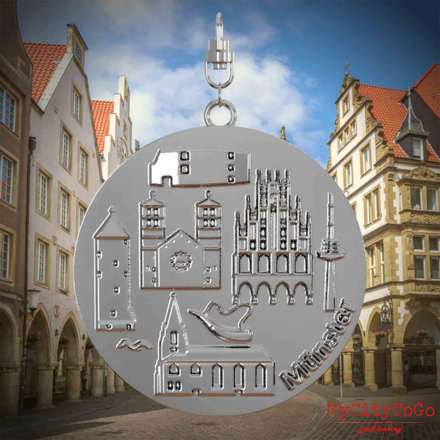 Halskette mit Drehwirbel und Vorderseite eines silbernen Schmuckanhängers, auf dem Reliefs bekannter Sehenswürdigkeiten der Stadt Muenster abgebildet sind. Den Hintergrund bildet eine Ansicht der Stadt Muenster.
