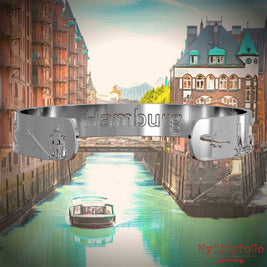 Inside of a silver bangle with reliefs of well-known landmarks of the city of Hamburg and an engraving of the city’s name. The background features a view of the city of Hamburg.