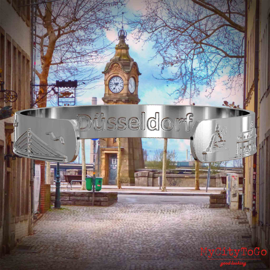 Innenseite eines silbernen Armreifs mit Reliefs bekannter Sehenswürdigkeiten der Stadt Duesseldorf und einer Gravur des Stadtnamens. Den Hintergrund bildet eine Ansicht der Stadt Duesseldorf.