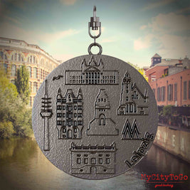 Halskette mit Drehwirbel und Vorderseite eines Schmuckanhängers aus Titan, auf dem Reliefs bekannter Sehenswürdigkeiten der Stadt Leipzig abgebildet sind. Den Hintergrund bildet eine Ansicht der Stadt Leipzig.
