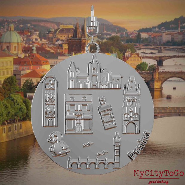 Halskette mit Drehwirbel und Vorderseite eines silbernen Schmuckanhängers, auf dem Reliefs bekannter Sehenswürdigkeiten der Stadt Prag abgebildet sind. Den Hintergrund bildet eine Ansicht der Stadt Prag.
