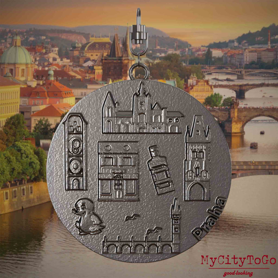 tHalskette mit Drehwirbel und Vorderseite eines Schmuckanhängers aus Titan, auf dem Reliefs bekannter Sehenswürdigkeiten der Stadt Prag abgebildet sind. Den Hintergrund bildet eine Ansicht der Stadt Prag.
