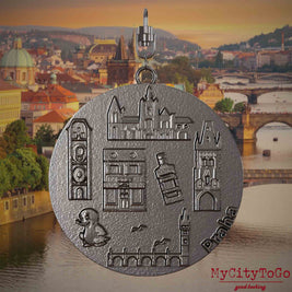 tHalskette mit Drehwirbel und Vorderseite eines Schmuckanhängers aus Titan, auf dem Reliefs bekannter Sehenswürdigkeiten der Stadt Prag abgebildet sind. Den Hintergrund bildet eine Ansicht der Stadt Prag.
