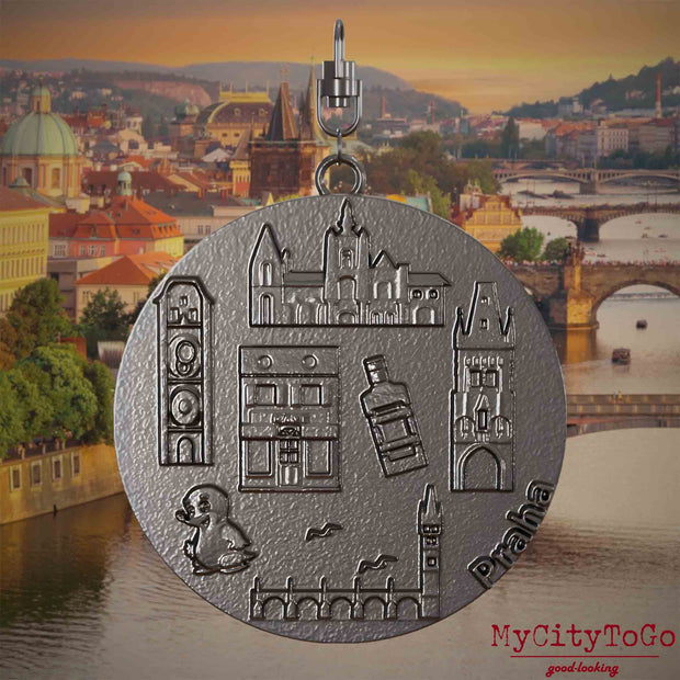 tHalskette mit Drehwirbel und Vorderseite eines Schmuckanhängers aus Titan, auf dem Reliefs bekannter Sehenswürdigkeiten der Stadt Prag abgebildet sind. Den Hintergrund bildet eine Ansicht der Stadt Prag.
