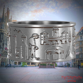 Ring aus Edelstahl mit Reliefs bekannter Sehenswürdigkeiten der Stadt Muenchen. Den Hintergrund bildet eine Ansicht der Stadt Muenchen.