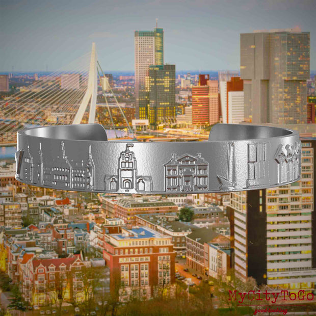 Armreif aus Edelstahl mit Reliefs bekannter Sehenswürdigkeiten der Stadt Rotterdam. Den Hintergrund bildet eine Ansicht der Stadt Rotterdam.