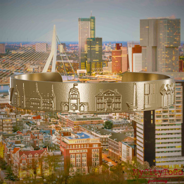 Armreif aus vergoldetem Edelstahl mit Reliefs bekannter Sehenswürdigkeiten der Stadt Rotterdam. Den Hintergrund bildet eine Ansicht der Stadt Rotterdam.