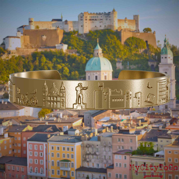 Armreif aus vergoldetem Edelstahl mit Reliefs bekannter Sehenswürdigkeiten der Stadt Salzburg. Den Hintergrund bildet eine Ansicht der Stadt Salzburg.