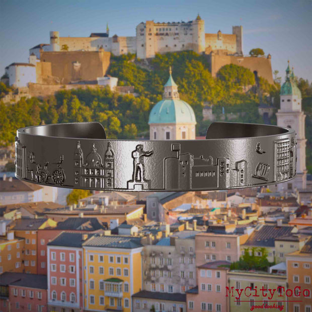 Armreif aus Titan mit Reliefs bekannter Sehenswürdigkeiten der Stadt Salzburg. Den Hintergrund bildet eine Ansicht der Stadt Salzburg.