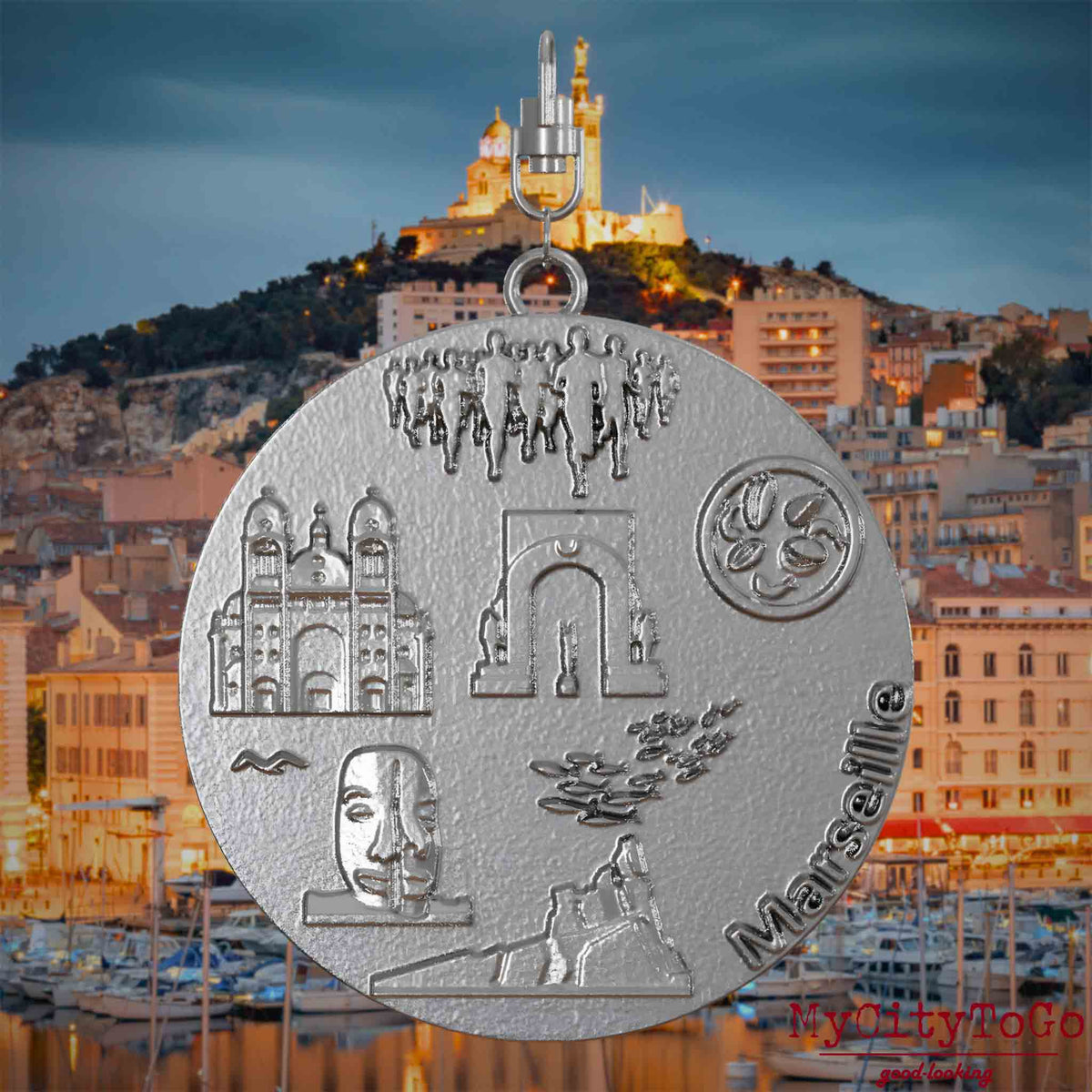 Halskette mit Drehwirbel und Rückseite eines Schmuckanhängers aus Edelstahl, auf dem Reliefs bekannter Sehenswürdigkeiten der Stadt Marseille abgebildet sind. Den Hintergrund bildet eine Ansicht der Stadt Marseille.
