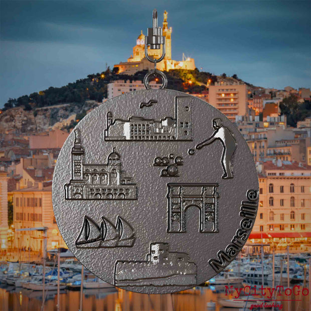Halskette mit Drehwirbel und Vorderseite eines Schmuckanhängers aus Titan, auf dem Reliefs bekannter Sehenswürdigkeiten der Stadt Marseille abgebildet sind. Den Hintergrund bildet eine Ansicht der Stadt Marseille.
