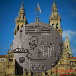 Halskette mit Drehwirbel und Rückseite eines Schmuckanhängers aus Titan, auf dem Reliefs bekannter Sehenswürdigkeiten des Jakobwegs abgebildet sind. Den Hintergrund bildet eine Ansicht der Stadt Santiago de Compostela.

