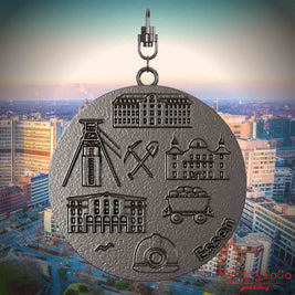 Halskette mit Drehwirbel und Rückseite eines Schmuckanhängers aus Titan, auf dem Reliefs bekannter Sehenswürdigkeiten der Stadt Essen abgebildet sind. Den Hintergrund bildet eine Ansicht der Stadt Essen.