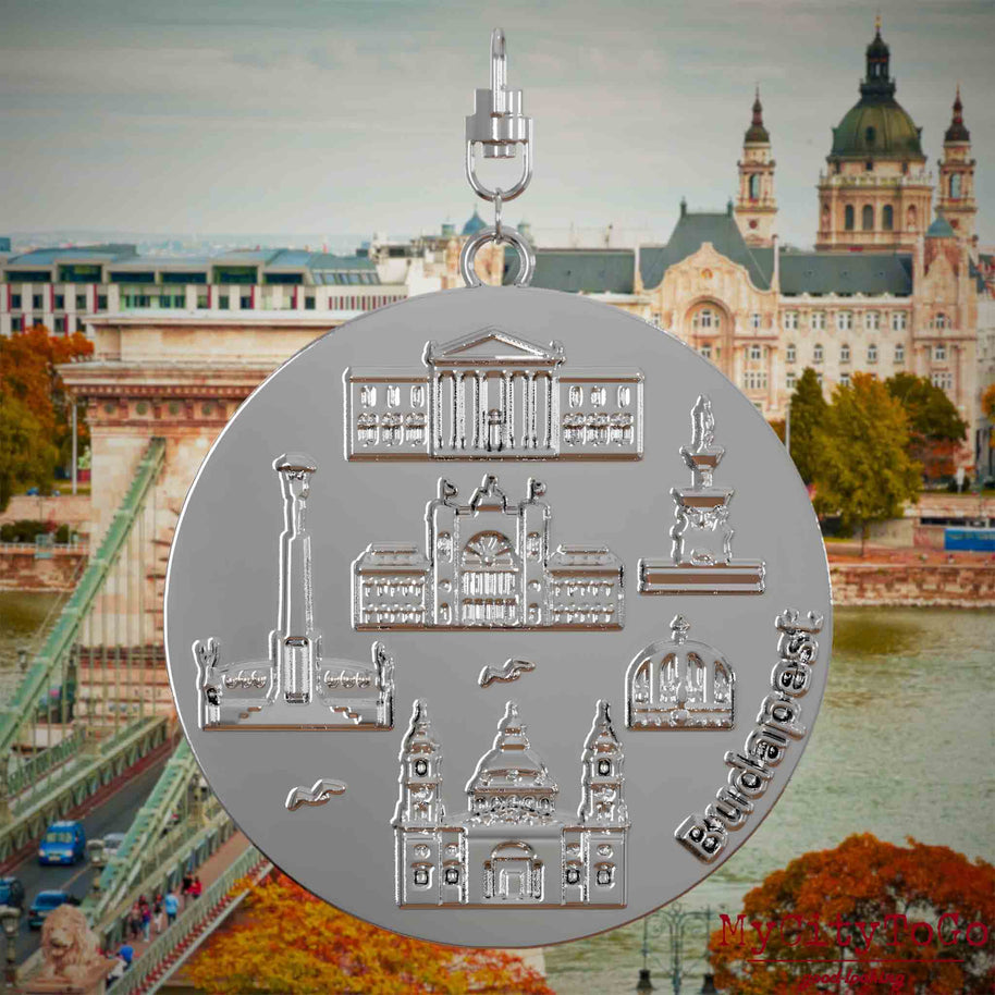 Halskette mit Drehwirbel und Rückseite eines silbernen Schmuckanhängers, auf dem Reliefs bekannter Sehenswürdigkeiten der Stadt Budapest abgebildet sind. Den Hintergrund bildet eine Ansicht der Stadt Budapest.

