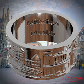 Versilberter Ring mit Reliefs bekannter Sehenswürdigkeiten der Stadt Muenchen und einer Gravur des Stadtnamens auf der Innenseite. Den Hintergrund bildet eine Ansicht der Stadt Muenchen.