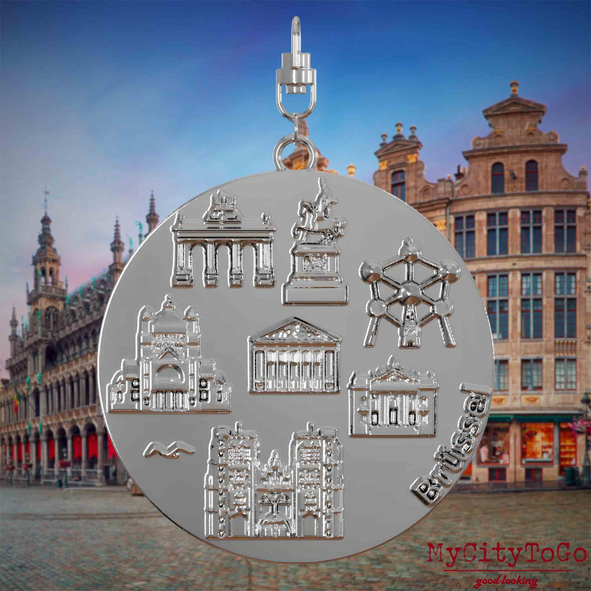 Halskette mit Drehwirbel und Rückseite eines silbernen Schmuckanhängers, auf dem Reliefs bekannter Sehenswürdigkeiten der Stadt Bruessel abgebildet sind. Den Hintergrund bildet eine Ansicht der Stadt Bruessel.
