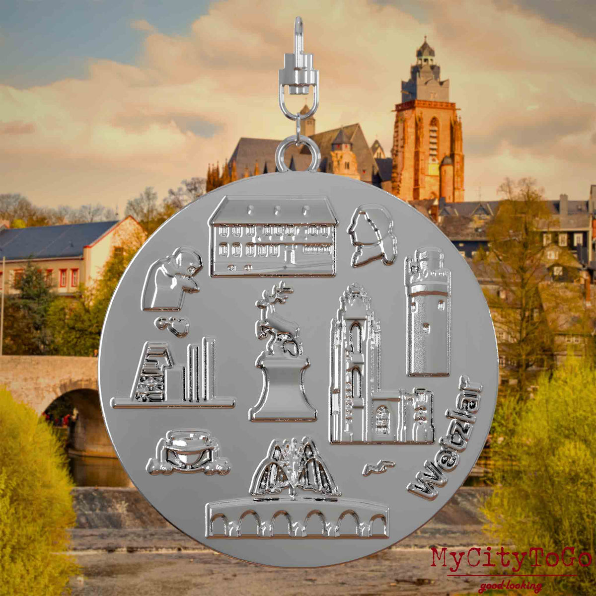 Halskette mit Drehwirbel und Rückseite eines silbernen Schmuckanhängers, auf dem Reliefs bekannter Sehenswürdigkeiten der Stadt Wetzlar abgebildet sind. Den Hintergrund bildet eine Ansicht der Stadt Wetzlar.

