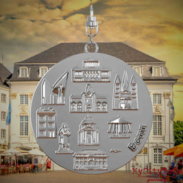 Halskette mit Drehwirbel und Vorderseite eines silbernen Schmuckanhängers, auf dem Reliefs bekannter Sehenswürdigkeiten der Stadt Bonn abgebildet sind. Den Hintergrund bildet eine Ansicht der Stadt Bonn.
