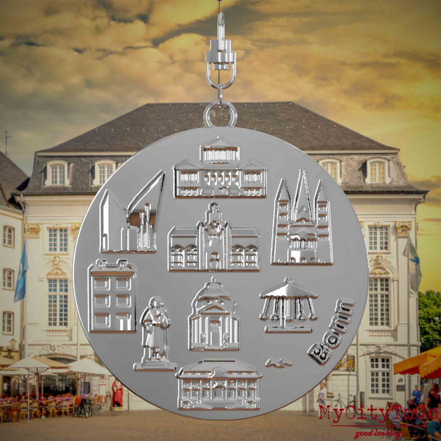 Halskette mit Drehwirbel und Vorderseite eines silbernen Schmuckanhängers, auf dem Reliefs bekannter Sehenswürdigkeiten der Stadt Bonn abgebildet sind. Den Hintergrund bildet eine Ansicht der Stadt Bonn.
