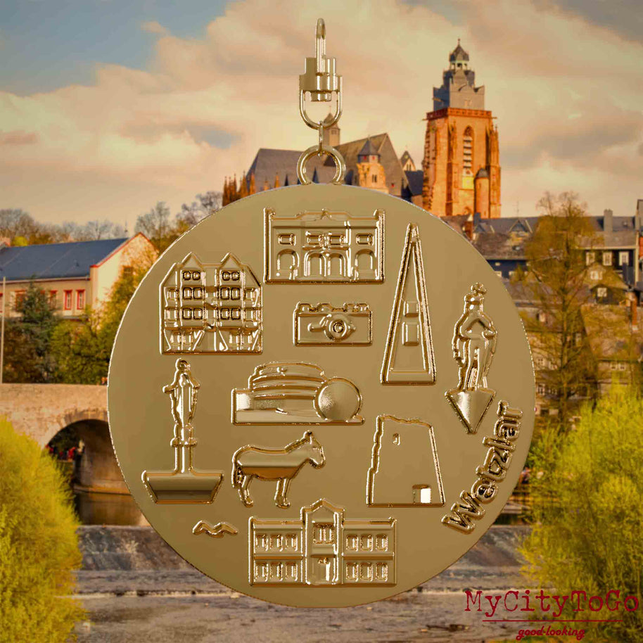 Halskette mit Drehwirbel und Vorderseite eines vergoldeten Schmuckanhängers, auf dem Reliefs bekannter Sehenswürdigkeiten der Stadt Wetzlar abgebildet sind. Den Hintergrund bildet eine Ansicht der Stadt Wetzlar.
