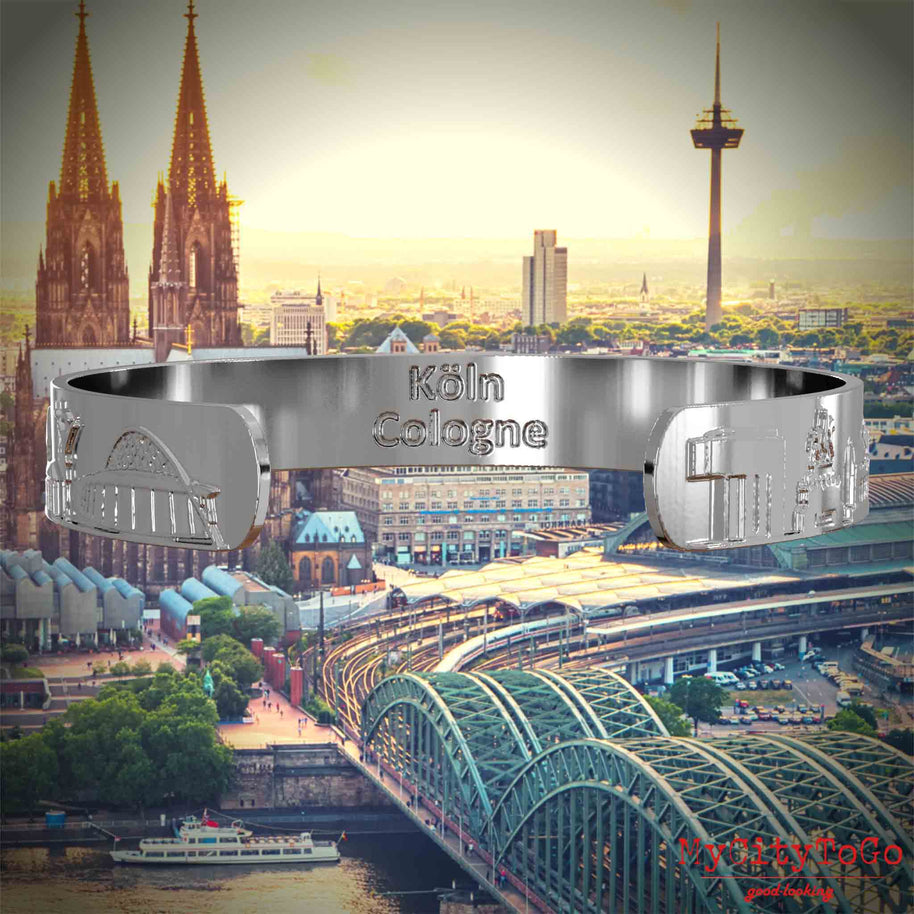 Innenseite eines silbernen Armreifs mit Reliefs bekannter Sehenswürdigkeiten der Stadt Köln und einer Gravur des Stadtnamens. Den Hintergrund bildet eine Ansicht der Stadt Koeln.