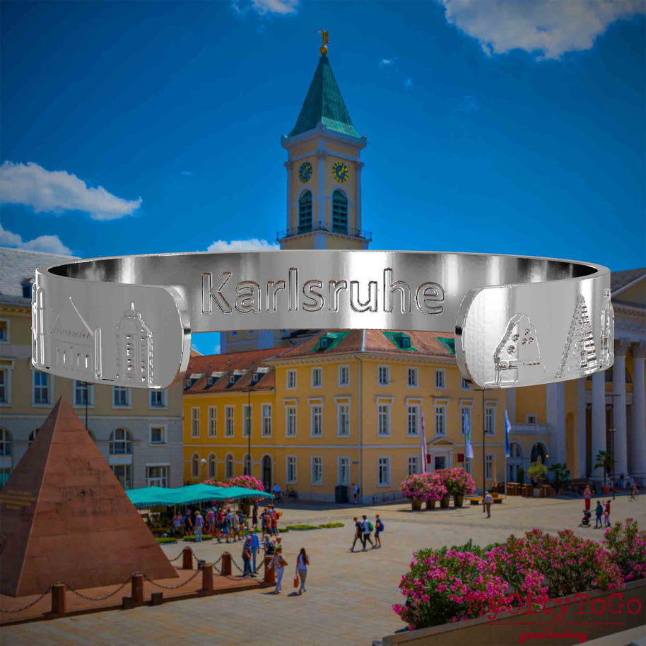 Innenseite eines silbernen Armreifs mit Reliefs bekannter Sehenswürdigkeiten der Stadt Karlsruhe und einer Gravur des Stadtnamens. Den Hintergrund bildet eine Ansicht der Stadt Karlsruhe.