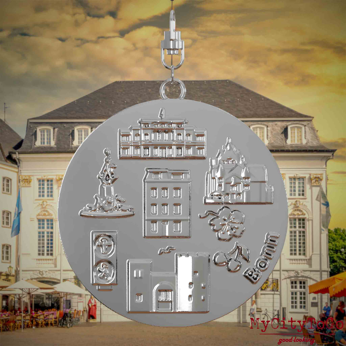 Halskette mit Drehwirbel und Rückseite eines silbernen Schmuckanhängers, auf dem Reliefs bekannter Sehenswürdigkeiten der Stadt Bonn abgebildet sind. Den Hintergrund bildet eine Ansicht der Stadt Bonn.
