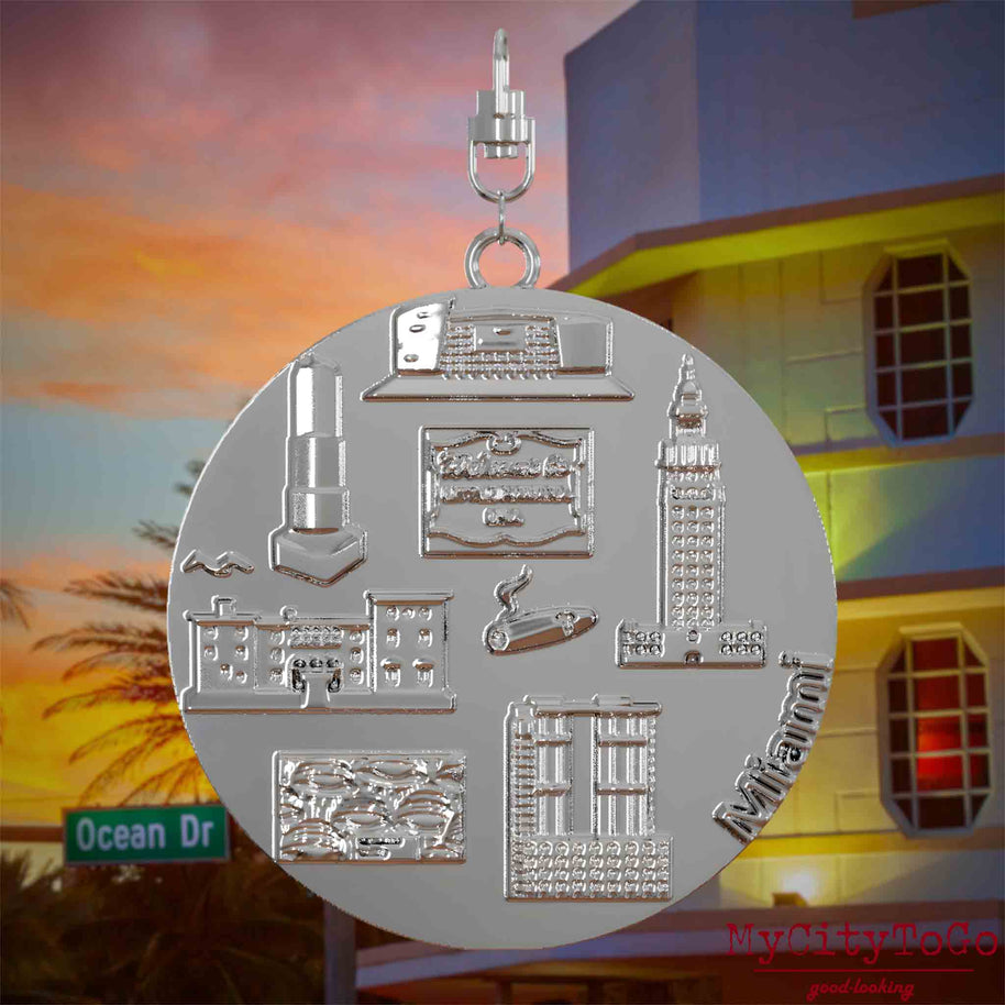 Halskette mit Drehwirbel und Vorderseite eines silbernen Schmuckanhängers, auf dem Reliefs bekannter Sehenswürdigkeiten der Stadt Miami abgebildet sind. Den Hintergrund bildet eine Ansicht der Stadt Miami.
