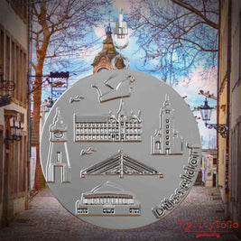 Halskette mit Drehwirbel und Rückseite eines silbernen Schmuckanhängers, auf dem Reliefs bekannter Sehenswürdigkeiten der Stadt Duesseldorf abgebildet sind. Den Hintergrund bildet eine Ansicht der Stadt Duesseldorf.