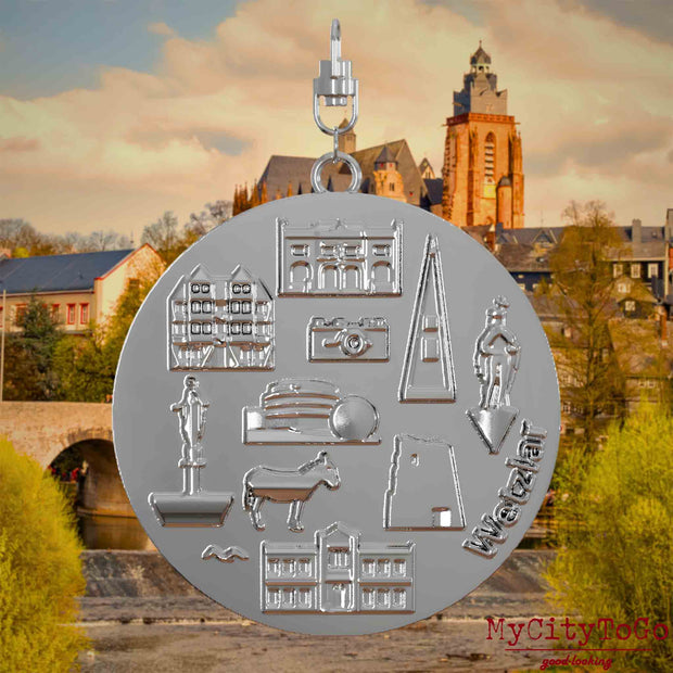 Halskette mit Drehwirbel und Vorderseite eines silbernen Schmuckanhängers, auf dem Reliefs bekannter Sehenswürdigkeiten der Stadt Wetzlar abgebildet sind. Den Hintergrund bildet eine Ansicht der Stadt Wetzlar.
