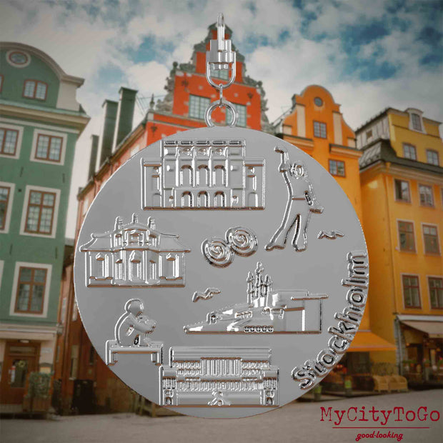 Halskette mit Drehwirbel und Vorderseite eines silbernen Schmuckanhängers, auf dem Reliefs bekannter Sehenswürdigkeiten der Stadt Stockholm abgebildet sind. Den Hintergrund bildet eine Ansicht der Stadt Stockholm.
