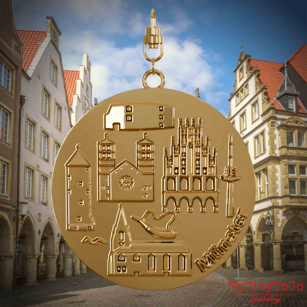 Halskette mit Drehwirbel und Vorderseite eines vergoldeten Schmuckanhängers, auf dem Reliefs bekannter Sehenswürdigkeiten der Stadt Muenster abgebildet sind. Den Hintergrund bildet eine Ansicht der Stadt Muenster.