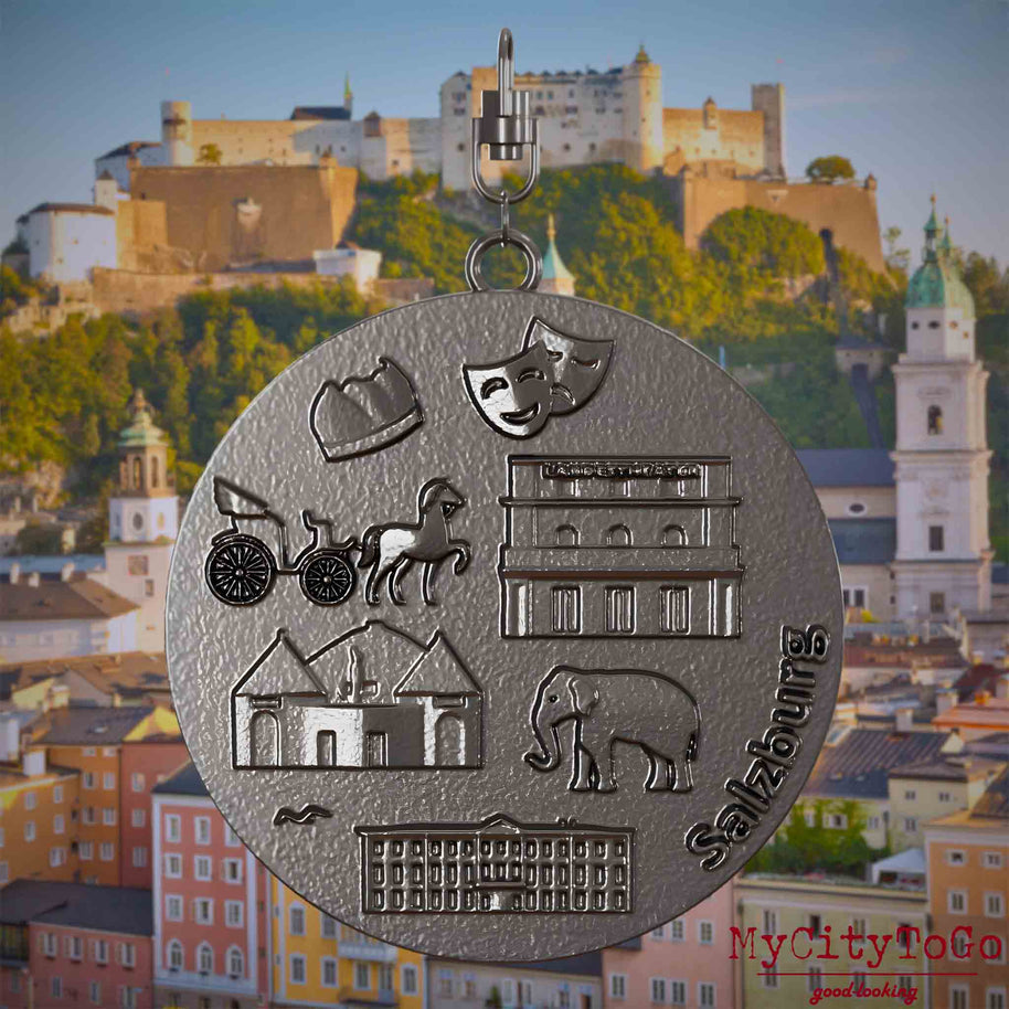 Halskette mit Drehwirbel und Rückseite eines Schmuckanhängers aus Titan, auf dem Reliefs bekannter Sehenswürdigkeiten der Stadt Salzburg abgebildet sind. Den Hintergrund bildet eine Ansicht der Stadt Salzburg.
