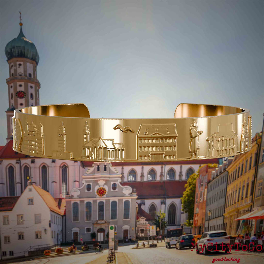 Vergoldeter Armreif mit Reliefs bekannter Sehenswürdigkeiten der Stadt Augsburg. Den Hintergrund bildet eine Ansicht der Stadt Augsburg.