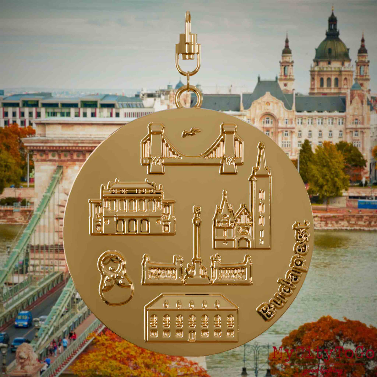 Halskette mit Drehwirbel und Vorderseite eines vergoldeten Schmuckanhängers, auf dem Reliefs bekannter Sehenswürdigkeiten der Stadt Budapest abgebildet sind. Den Hintergrund bildet eine Ansicht der Stadt Budapest.
