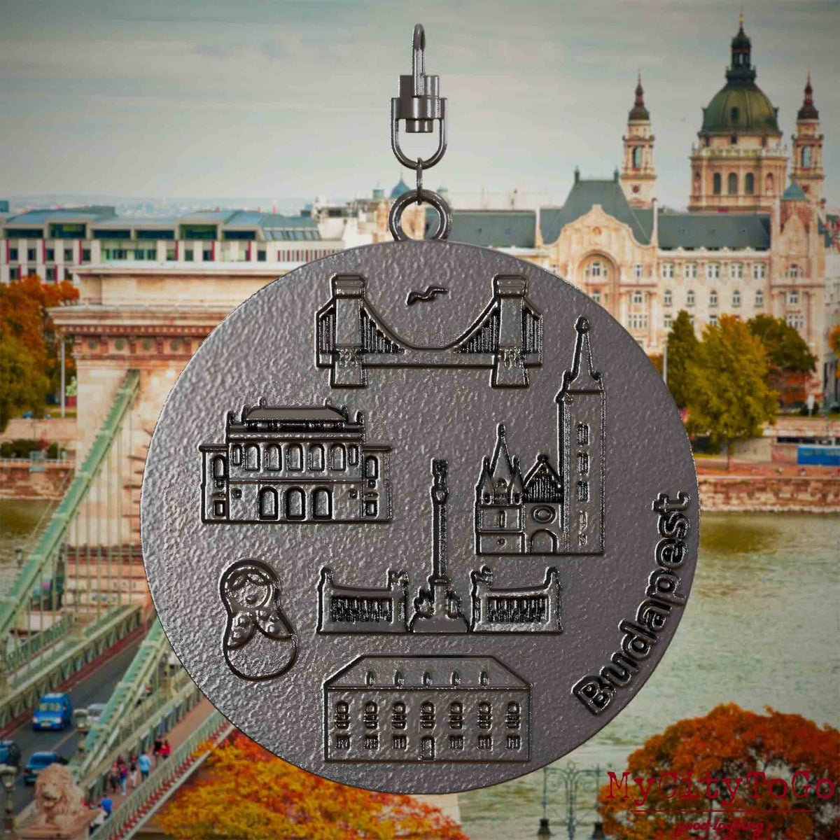 Halskette mit Drehwirbel und Vorderseite eines Schmuckanhängers aus Titan, auf dem Reliefs bekannter Sehenswürdigkeiten der Stadt Budapest abgebildet sind. Den Hintergrund bildet eine Ansicht der Stadt Budapest.
