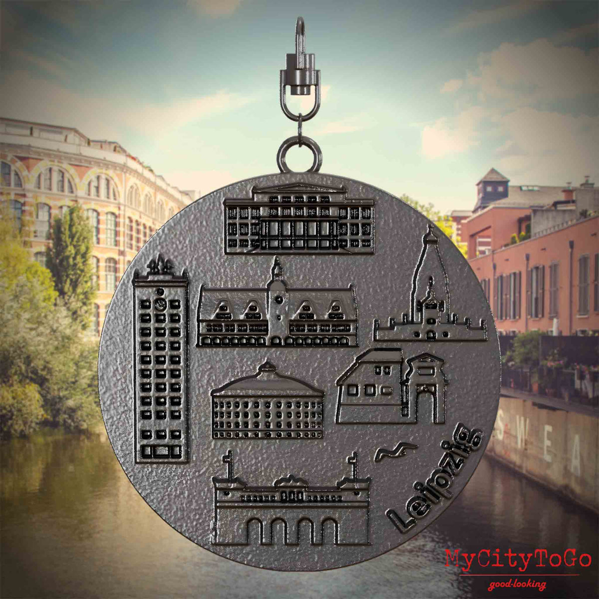 Halskette mit Drehwirbel und Rückseite eines Schmuckanhängers aus Titan, auf dem Reliefs bekannter Sehenswürdigkeiten der Stadt Leipzig abgebildet sind. Den Hintergrund bildet eine Ansicht der Stadt Leipzig.