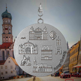 Halskette mit Drehwirbel und Rückseite eines Schmuckanhängers aus Edelstahl, auf dem Reliefs bekannter Sehenswürdigkeiten der Stadt Augsburg abgebildet sind. Den Hintergrund bildet eine Ansicht der Stadt Augsburg.
