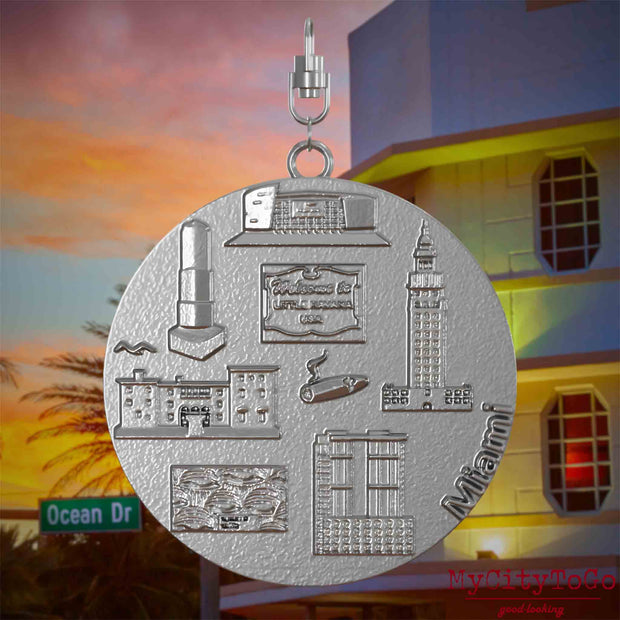 Halskette mit Drehwirbel und Vorderseite eines Schmuckanhängers aus Edelstahl, auf dem Reliefs bekannter Sehenswürdigkeiten der Stadt Miami abgebildet sind. Den Hintergrund bildet eine Ansicht der Stadt Miami.
