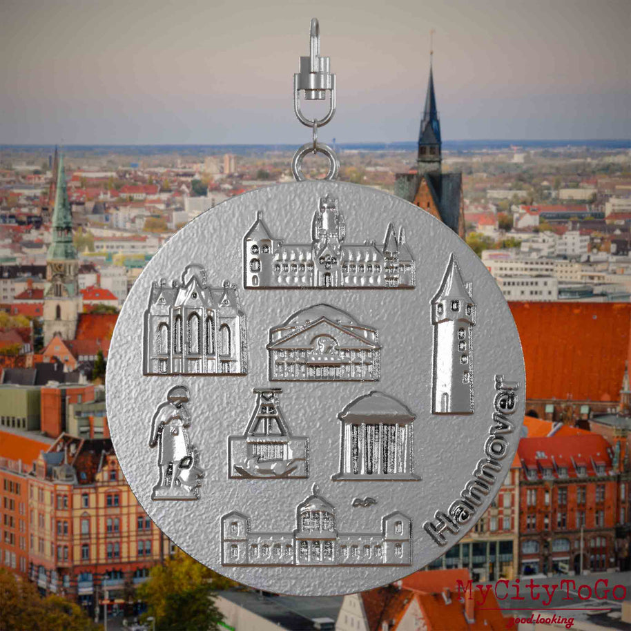 Halskette mit Drehwirbel und Vorderseite eines Schmuckanhängers aus Edelstahl, auf dem Reliefs bekannter Sehenswürdigkeiten der Stadt Hannover abgebildet sind. Den Hintergrund bildet eine Ansicht der Stadt Hannover.
