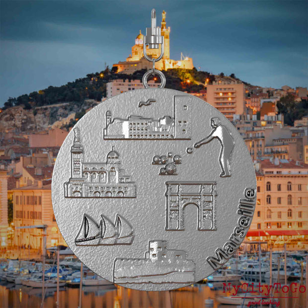 Halskette mit Drehwirbel und Vorderseite eines Schmuckanhängers aus Edelstahl, auf dem Reliefs bekannter Sehenswürdigkeiten der Stadt Marseille abgebildet sind. Den Hintergrund bildet eine Ansicht der Stadt Marseille.

