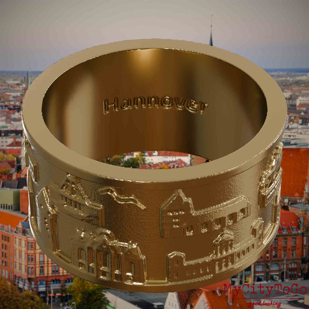 Ring aus vergoldetem Edelstahl mit Reliefs bekannter Sehenswürdigkeiten der Stadt Hannover und einer Gravur des Stadtnamens auf der Innenseite. Den Hintergrund bildet eine Ansicht der Stadt Hannover.