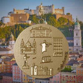 Halskette mit Drehwirbel und Vorderseite eines Schmuckanhängers aus vergoldetem Edelstahl, auf dem Reliefs bekannter Sehenswürdigkeiten der Stadt Salzburg abgebildet sind. Den Hintergrund bildet eine Ansicht der Stadt Salzburg.
