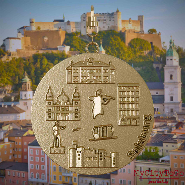 Halskette mit Drehwirbel und Vorderseite eines Schmuckanhängers aus vergoldetem Edelstahl, auf dem Reliefs bekannter Sehenswürdigkeiten der Stadt Salzburg abgebildet sind. Den Hintergrund bildet eine Ansicht der Stadt Salzburg.
