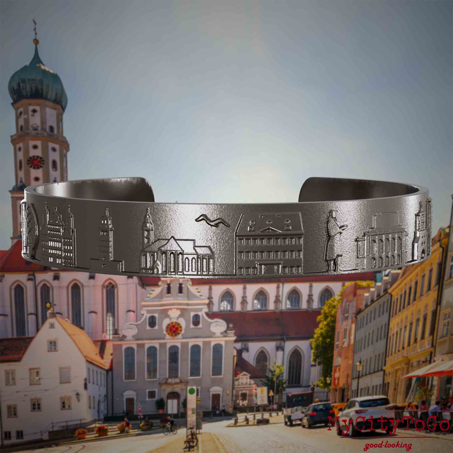 Armreif aus Titan mit Reliefs bekannter Sehenswürdigkeiten der Stadt Augsburg. Den Hintergrund bildet eine Ansicht der Stadt Augsburg.