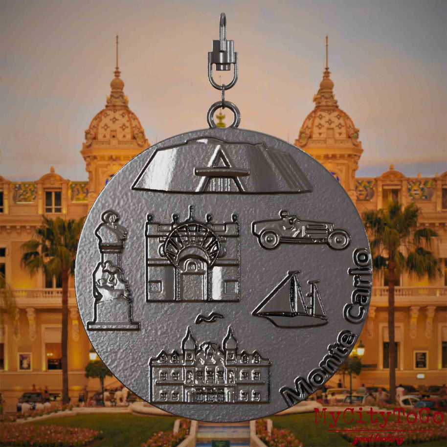 Halskette mit Drehwirbel und Vorderseite eines Schmuckanhängers aus Titan, auf dem Reliefs bekannter Sehenswürdigkeiten der Stadt Monte Carlo abgebildet sind. Den Hintergrund bildet eine Ansicht der Stadt Monte Carlo.
