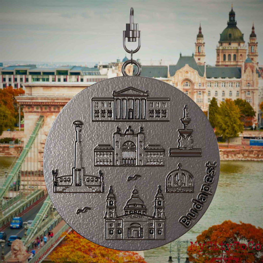 Halskette mit Drehwirbel und Rückseite eines Schmuckanhängers aus Titan, auf dem Reliefs bekannter Sehenswürdigkeiten der Stadt Budapest abgebildet sind. Den Hintergrund bildet eine Ansicht der Stadt Budapest.
